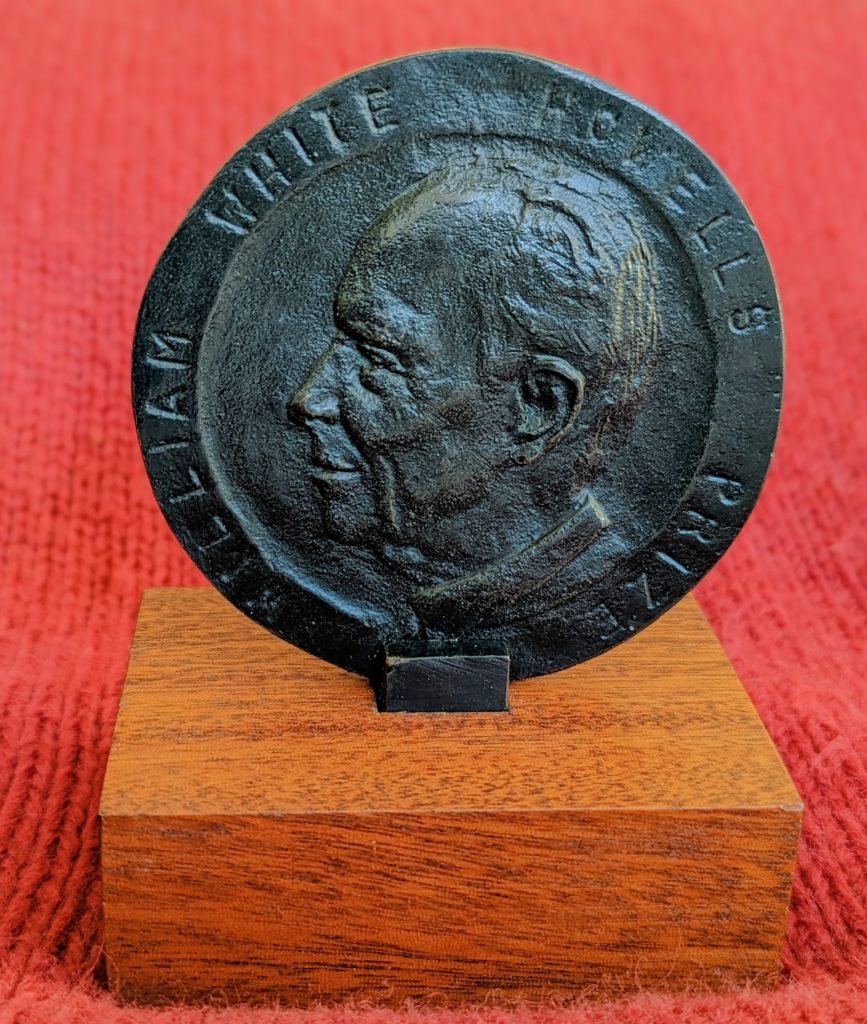 photo of the medal. It is a dark grey/black medal, with the profile of a man in the center. Around the edge it says "William White Howells Prize". The medal is sitting on a wood block with a red knit background under and behind it.